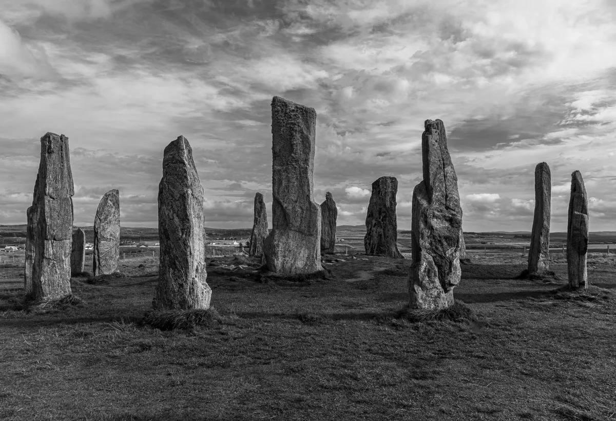 Conjunto de piedras verticales sobre hierba bajo un cielo de nubes, fotografiado en blanco y negro.